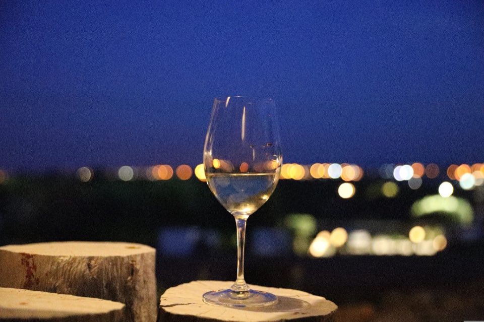A stemmed glass of pale wine on a wooden stump at night, with distant city lights