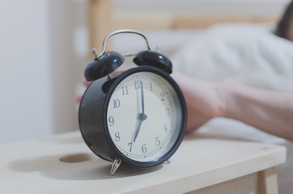 Black twin-bell alarm clock showing about 7:00 beside a person reaching to turn it off