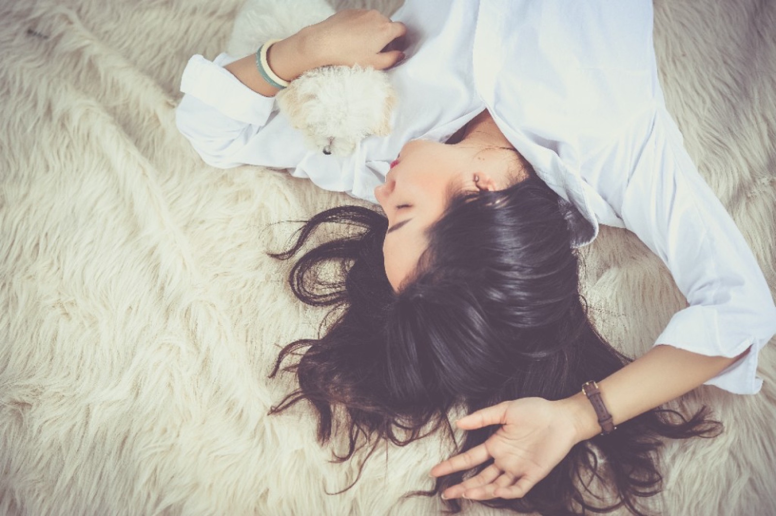 A woman asleep on a soft rug with a small white puppy, a watch on her wrist