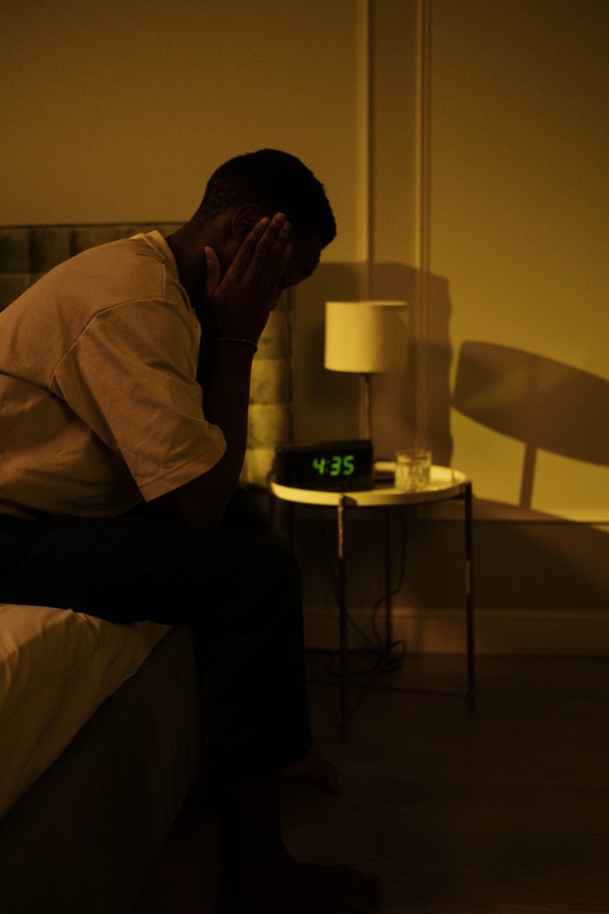 A person sitting on the edge of a bed at night, face in their hands, beside a dim lamp and a digital clock reading 4:35